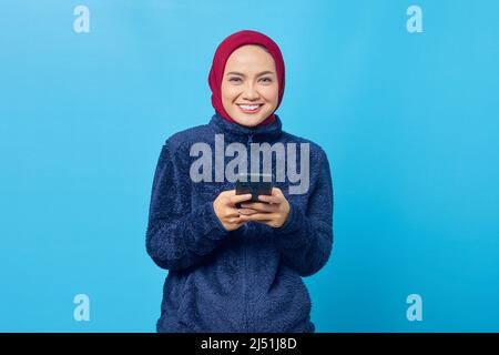 Smilling young Asian woman using smartphone and looking at camera on blue background Stock Photo