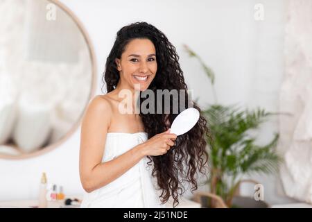 Glad young caucasian brunette woman in towel combing long curly hair with comb in light bedroom interior Stock Photo