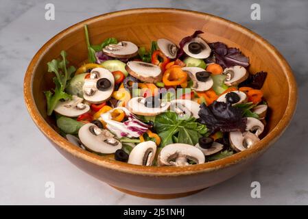 Organic mixed greens, mushrooms and peppers salad served garnished with black olive in old, used wooden bowl on marble table Stock Photo