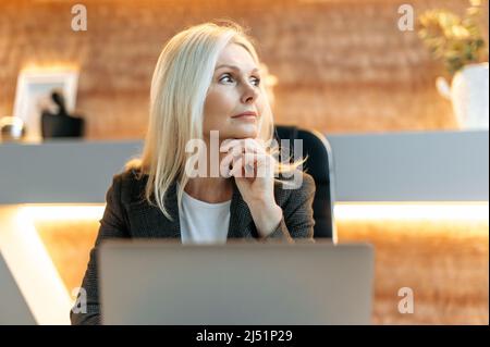 Pensive middle-aged blonde woman, ceo, manager, broker, banker sits at a table in modern office, dressed in formal suit, thinks over a business idea or financial strategy, planning, looks to the side Stock Photo