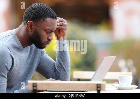 Profile of a worried man with black skin checking laptop in a bar terrace Stock Photo
