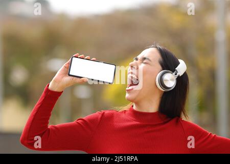 Happy woman in red wearing headphones singing in the street with blank smart phone screen Stock Photo