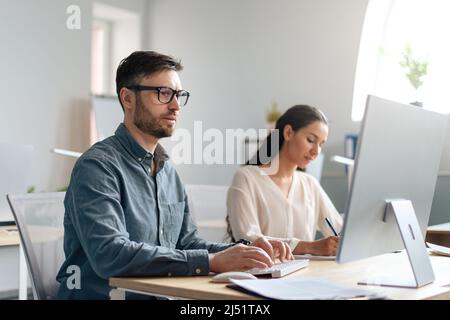 Focused Caucasian man using computer, working in modern office with his colleague, copy space Stock Photo