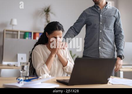 Upset young woman crying near laptop pc at office, male colleague helping and comforting her Stock Photo