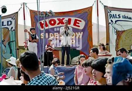 An announcer or barker with a microphone addressing the passing crowds outside the Hoxie Brothers Sideshow and Combined Zoo at its travelling circus in the USA in 1965. Alongside him is a boy holding knives and a young man with two clown puppets. Behind are colourful hand-painted banners advertising the attractions. This image is from an old American amateur (badly damaged) colour transparency. It has been heavily retouched and will look its best used at small sizes – a vintage 1960s photograph. Stock Photo