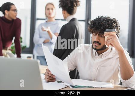 Tired indian businessman holding coffee and document near laptop in office Stock Photo