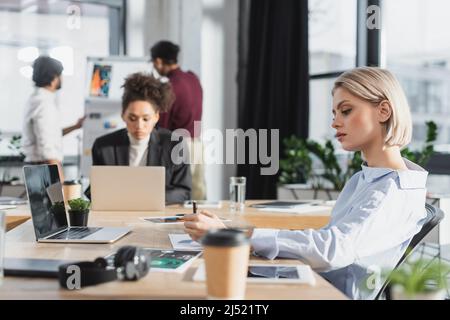 Young businesswoman looking at documents near coffee to go and laptop in office Stock Photo