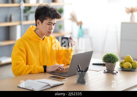 Focused asian man watching video on pc, drinking hot coffee Stock Photo