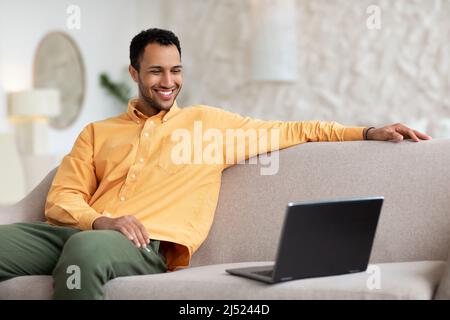 Smiling young man watching video on computer, sitting on couch Stock Photo