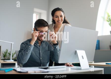 Two happy colleagues working on startup project together, discussing ideas, using modern computer at open space office Stock Photo