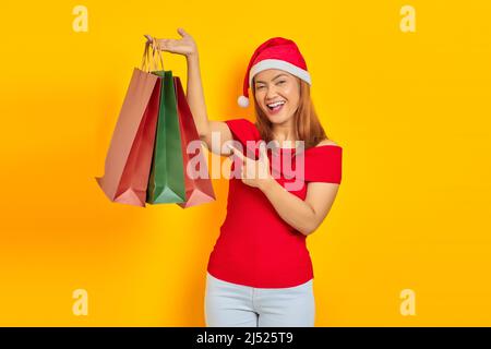 Cheerful young Asian woman in Santa Claus hat pointing finger at shopping bag on yellow background Stock Photo