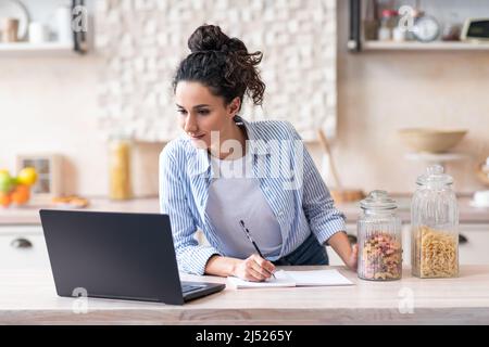 Latin woman writing new recipe watching online lessons on laptop in light kitchen interior, copy space Stock Photo