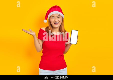 Smiling young Asian woman in Santa Claus hat extending hand inviting to come and showing smartphone on yellow background Stock Photo