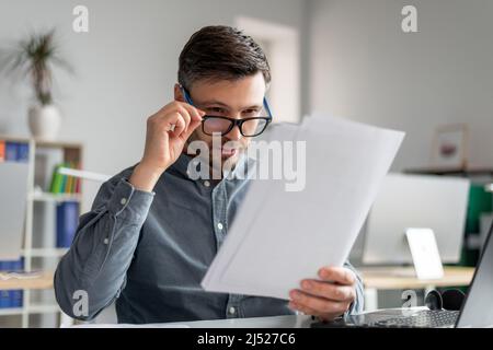 Concerned mature businessman in eyeglasses reading documents at workplace in office, checking annual reports Stock Photo