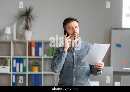 Serious middle aged businessman working at office, standing by workdesk, holding documents and having phone conversation Stock Photo