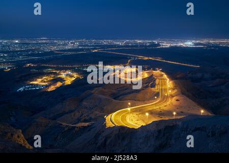 Winding road in mountains against illuminated city at night. Al Ain in Abu Dhabi Emirate. Stock Photo