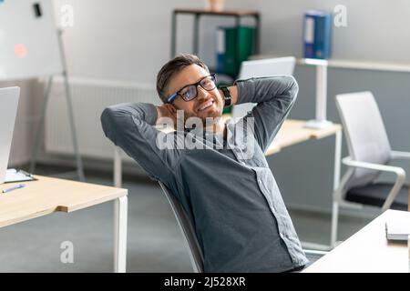 Confident middle aged company employee leaning on chair with hands behind head, relaxing and smiling at camera in office Stock Photo