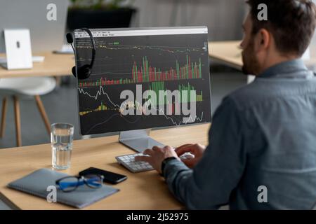 Unrecognizable mature businessman sitting at workdesk in office and typing on keyboard with graph on computer monitor Stock Photo