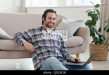 Cheerful millennial european guy with stubble blogger or freelancer in shirt working on laptop in living room Stock Photo