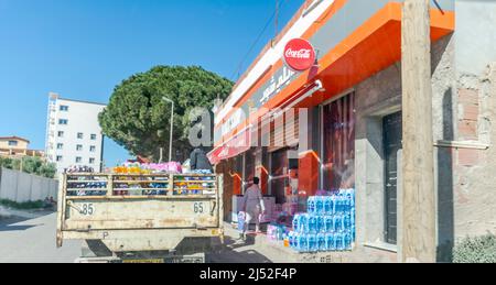 Mineral water bottles stocked outside under sunlight, a truck loading juice and soda packs is parked, two unrecognizable men unloading on the sidewalk Stock Photo