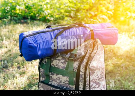 Defocus military backpack and blue tent or sleeping bag. Army bag on green grass background near tree. Military camouflage army rucksack. Tourist Stock Photo