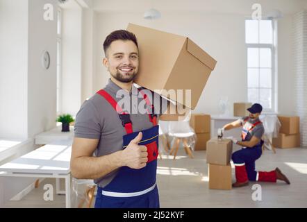 Smiling male worker of moving and delivery company holding cardboard box showing thumbs up. Stock Photo
