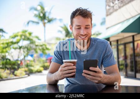Phone app young man using technology device holding cellphone at cafe texting with messaging sms apps drinking coffee in summer. Handsome young casual Stock Photo