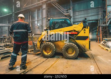 Workshop of metallurgical plant with small loading tractor or mower and unrecognizable engineer. Stock Photo