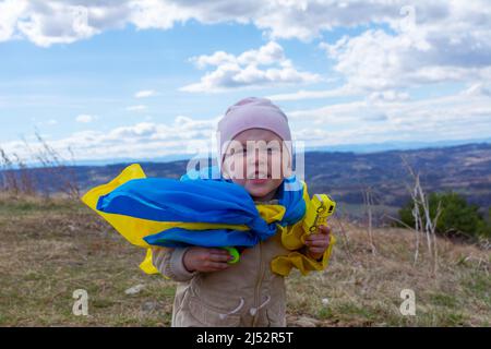 A baby girl with a flag of Ukraine against the hills and Ukrainian nature and blue cloudy sky. Portrait of a patriotic baby girl with the flag of Ukra Stock Photo