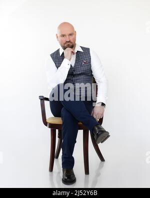 Portrait of thoughtful middle-aged businessman in grey vest, blue jeans, white shirt sitting, putting head on chin. Stock Photo