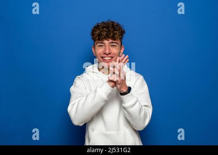 Caucasian young man with curly hair dressed in a white sweatshirt clapping hands smiling, isolated on blue studio background Stock Photo