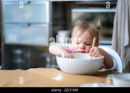 Little cute funny girl licking the dough from her finger helping mother prepare pie cake in kitchen, baking homemade cookie together. Stock Photo