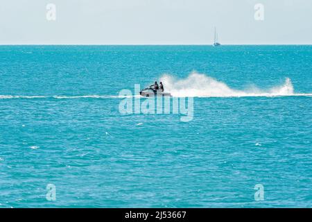 Airlie Beach, Whitsundays, Queensland, Australia - April 2022: Two people racing across the ocean on a jet ski with a sailing yacht in the background Stock Photo