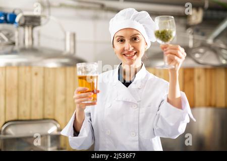 Woman brewer holding glass of hops and glass of beer in her hands at brewery Stock Photo