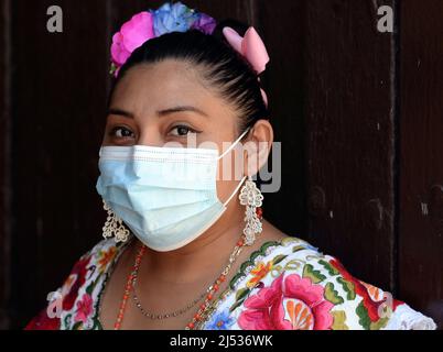 Body-positive young Mexican woman with flowers in slicked-back hair wears a colorful traditional Yucatan Maya huipil dress and surgical face mask. Stock Photo