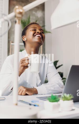 African woman drinking tea during lunch break and using headphones to talk to her friends with whom she is having fun and laughing. Lady boss. Modern Stock Photo