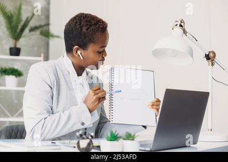 African-american woman teacher sitting at home giving online maths lessons using laptop. Math teacher showing notebook at web camera . Video Stock Photo