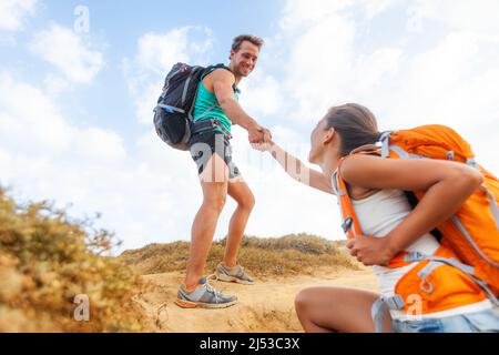People hiking man helping woman climbing up hill on mountain hike. Helping hand team support friend supporting success trek for girl Stock Photo