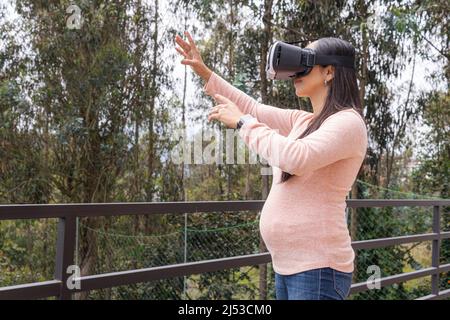 Pregnant Latin woman touching something using virtual reality glasses in the middle of a forest on a terrace Stock Photo