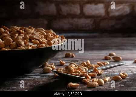 Roasted peanuts on a black bowl and on a spoon over a rustic wooden table with a brick wall in the background Stock Photo