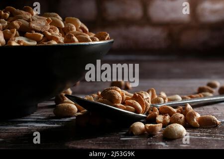 Roasted peanuts on a black bowl and on a spoon over a rustic wooden table with a brick wall in the background Stock Photo