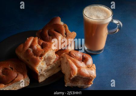 Cinnamon bread or rolls on a black plate  with a glass cup of hot coffee cocoa chocolate moka cappuccino, on a dark blue kitchen table, side view Stock Photo