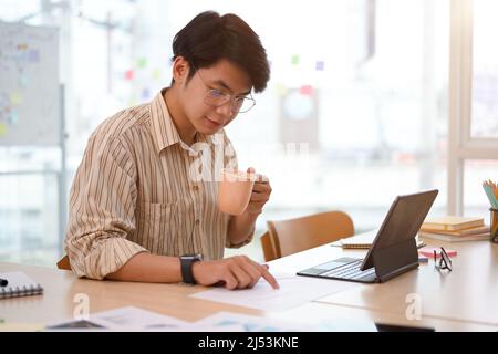 Focused Asian businessman or male entrepreneur sipping coffee while checking sale and stock report in the office. Stock Photo