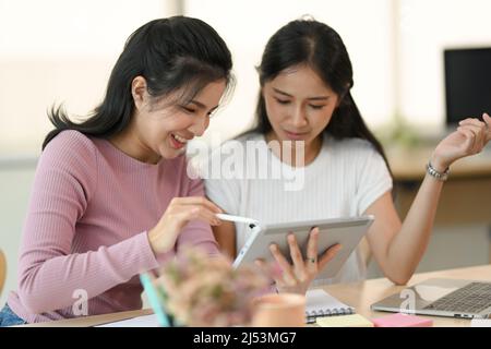 Two Asian female colleagues friends are working together, using tablet touchpad and looking something at screen in the office. Stock Photo