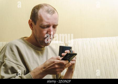 A white man sits on a couch and reads the news on his smartphone. Portrait of a man with a phone and a black cup Stock Photo
