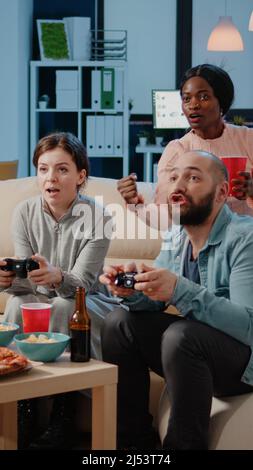 Diverse group of workmates using joysticks to play video games on television. Colleagues cheering while playing game with controllers on tv console to have fun and entertainment. Stock Photo