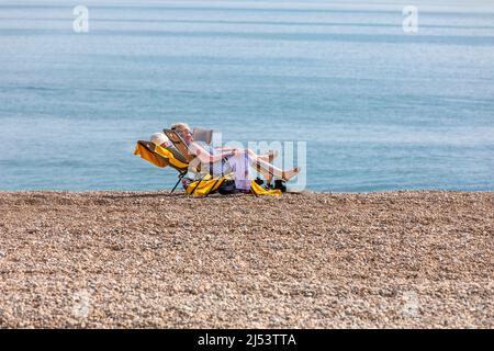 A couple sunbathing in folding chairs on a shingle beach by a calm blue sea Stock Photo