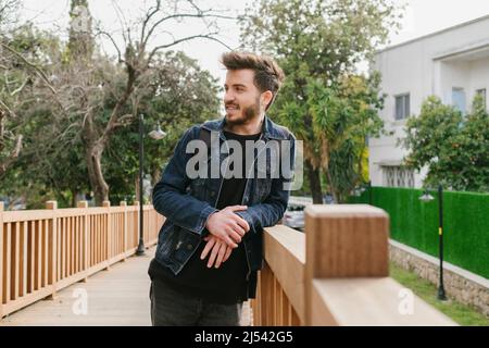 Portrait of young handsome man smiling outdoor. Young man wearing denim clothes looking away with big smile while leaning to wooden fence at city park Stock Photo