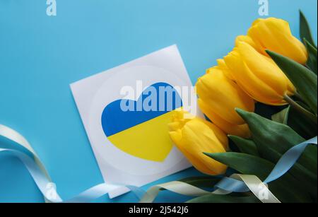 Сard with a heart in the colors of the Ukrainian flag and yellow tulips on a blue background. Support for Ukraine Stock Photo
