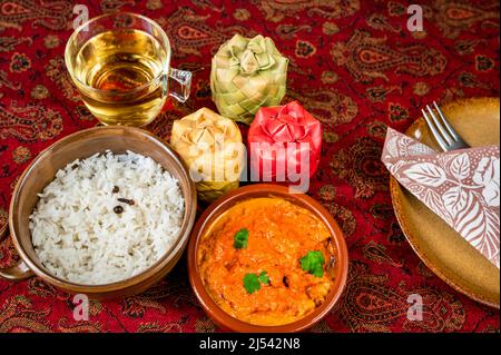 Chicken tikka masala, stewed rice in 2 bowl,cup of tea, 3 bamboo spice container, part of plate, towel and fork on red tablecloth. Indian food. Stock Photo